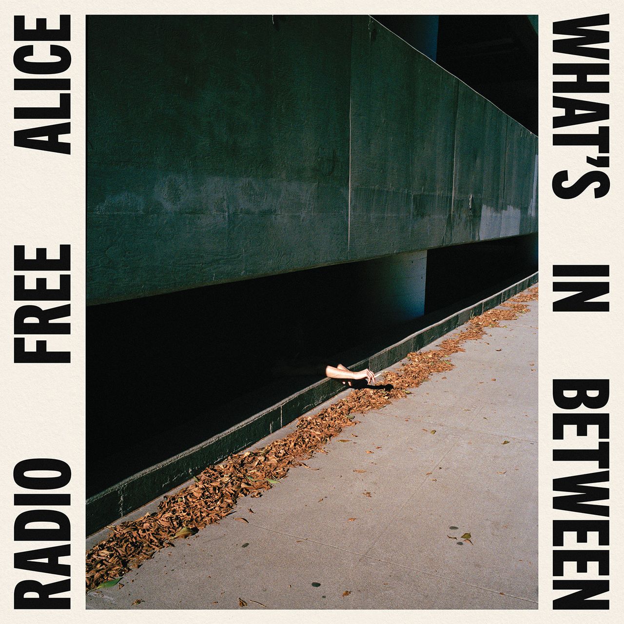Album cover for Radio Free Alice's 'What's In Between', showing hands reaching out from a narrow concrete gap beside a leaf-strewn path.